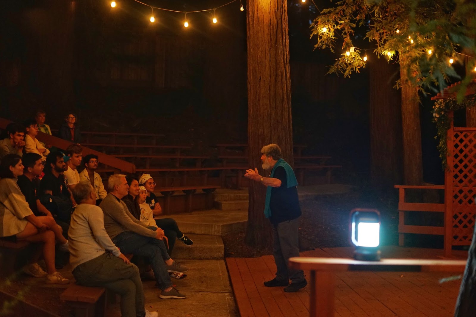 Mr. Joseph Lurie, animated, in the light of a portable lamp, reciting stories to everyone at the retreat. The setting is an amphitheater amidst redwoods.