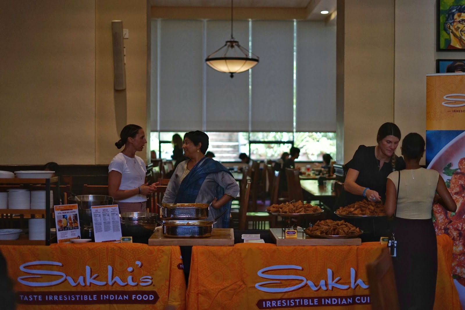 A pair of tables is set up in the dining hall, on which the cloth on the table, reads ‘Sukhi’s, Taste Irresistible Indian Food. Placards showcasing the list of products offered by Sukhi’s rests on the table. Sanjog, the owner stands animated speaking to her associate. There is another associate in white on the right, serving a sample of the samosa and chutney to a resident