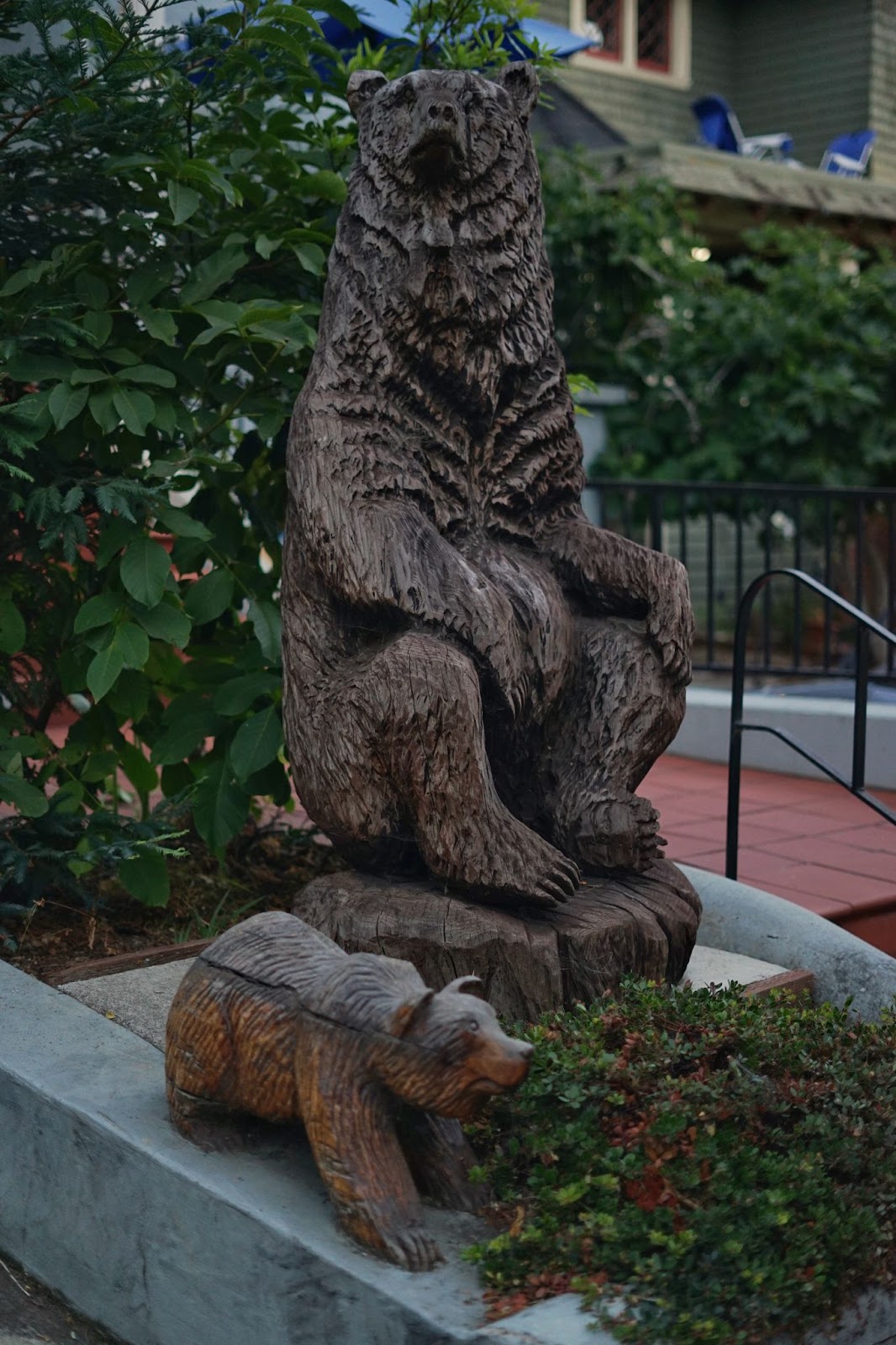 A wood carved statue of a bear sitting upright, with a small bear cub carving walking next to it. 