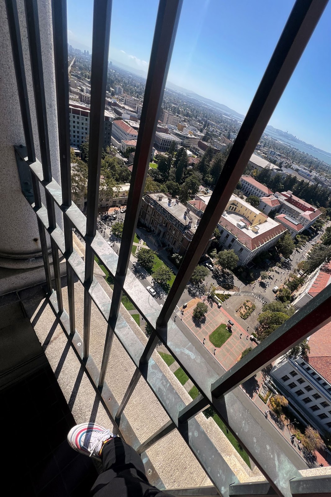 A first-person view looking down at shoes. The person is atop. a railing in Sather Tower.