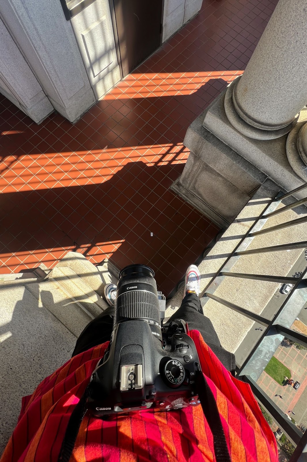 A first-person view looking down at a camera and shoes. The person is atop. a railing in Sather Tower.