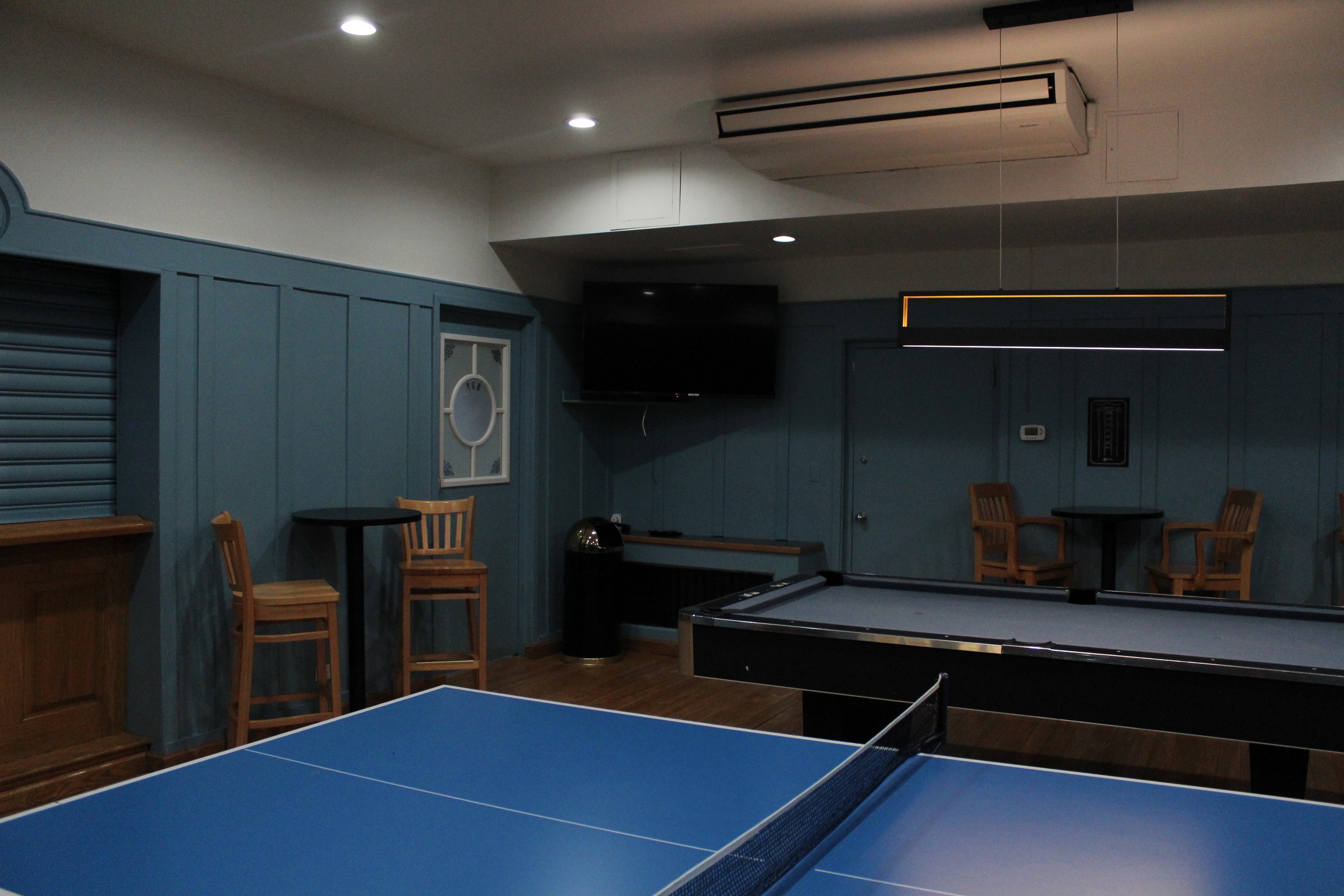 A game room with blue wood-panelled walls and high chairs and pool equipment.
