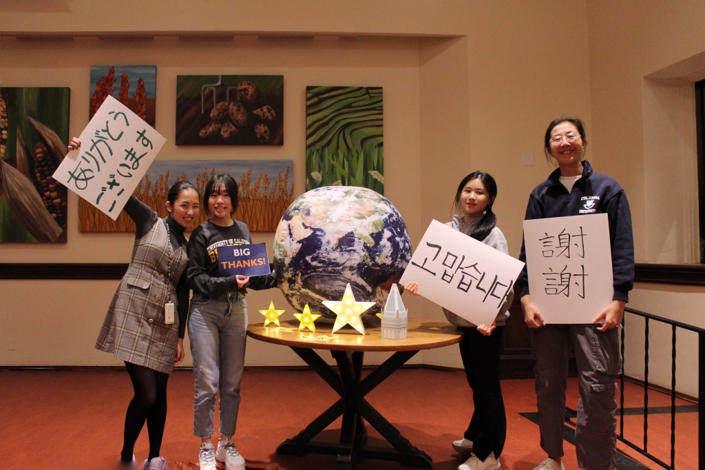 Residents hold up thanks in various languages near a table adorned with a globe and Big Give decorations.